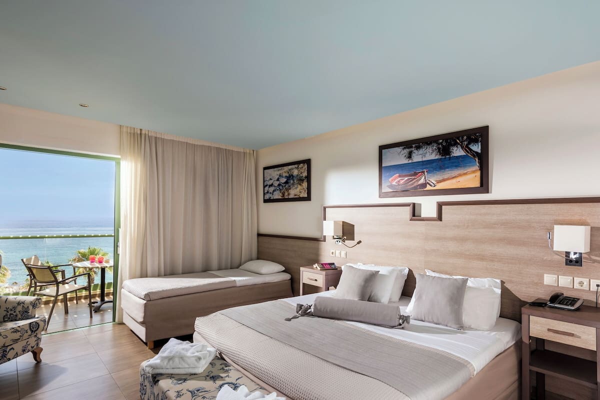 A hotel room with a bed, bedside tables, a chair, a small table, and a view of the sea.