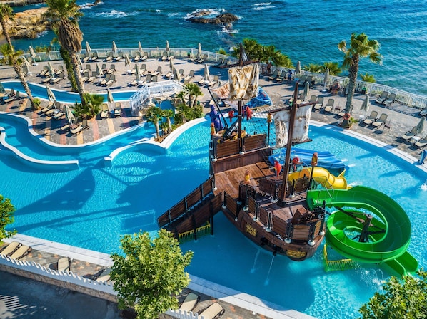 Una gran piscina al aire libre con una zona de juegos con forma de barco pirata, rodeada de sillas de descanso y palmeras.