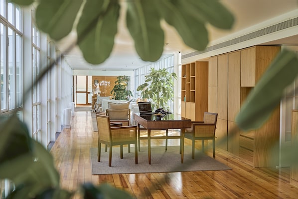 A modern interior with a wooden dining table, chairs, and a laptop. There are large windows and a plant in the background.