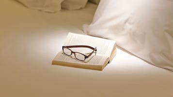 Um livro com óculos por cima, sobre uma cama com lençóis e almofadas brancas.