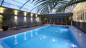 Une piscine intérieure dotée de chaises longues et de plantes en pot, entourée de murs de verre et surmontée d’un plafond haut.