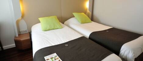 Habitación de hotel con dos camas, cada una con almohada y manta. Se observa una mesita de noche con lámpara y un pequeño reloj.