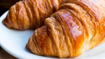 Mesa de café da manhã com croissants em um prato, um copo de suco de laranja e uma xícara ao fundo.
