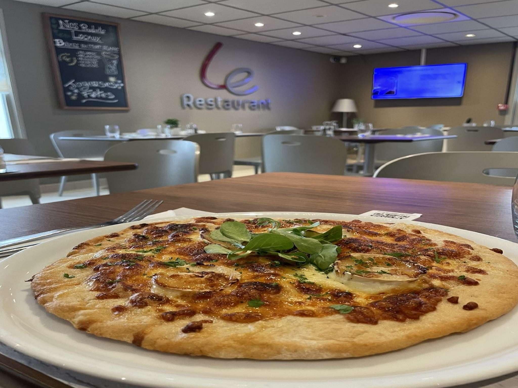 Una pizza sobre un plato blanco en un restaurante con un menú en una pizarra y una pantalla de televisión al fondo.