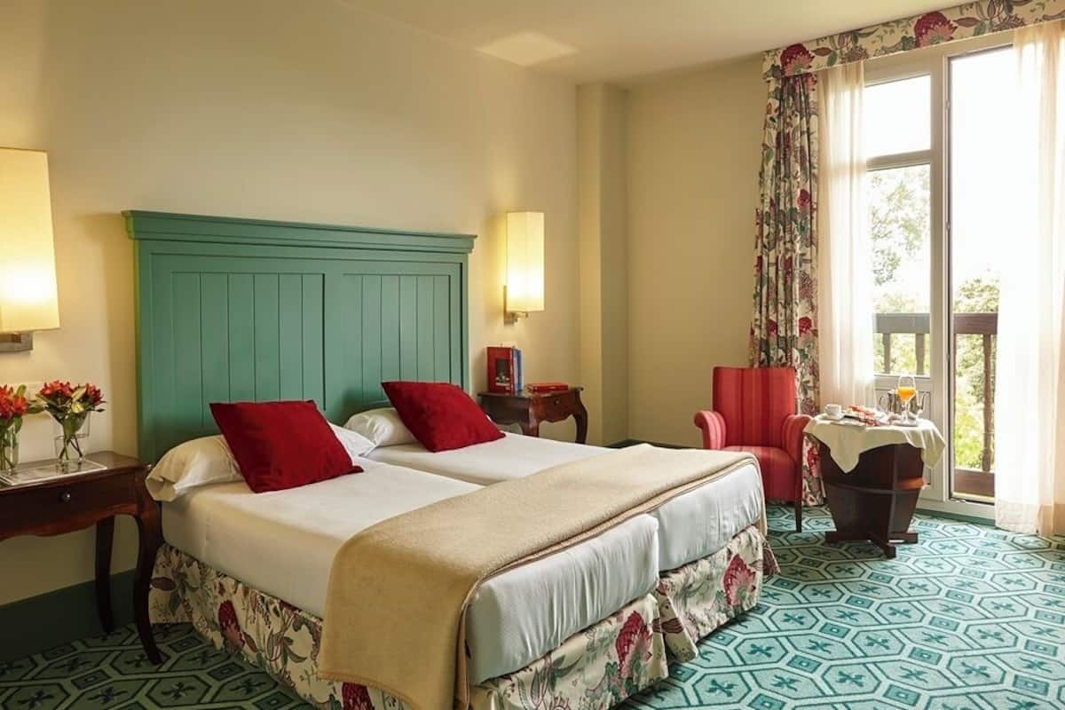 Habitación de hotel con dos camas, una silla roja, una mesita con una bebida y un balcón con vistas.