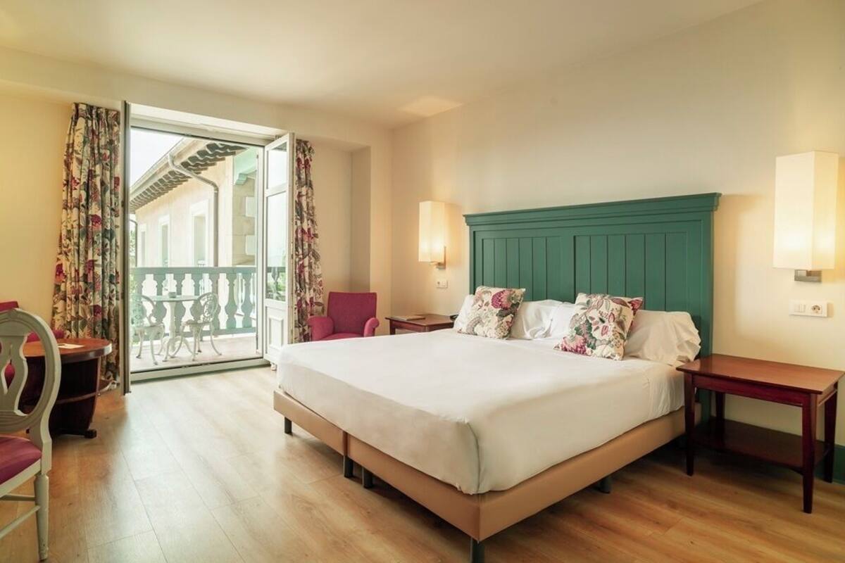 Habitación de hotel con cama, una silla, una mesa y un balcón que ofrece vistas a edificios.