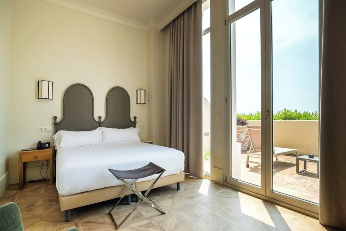 Habitación de hotel con una cama grande, una silla, una mesita de noche y vistas del patio exterior a través de una puerta corrediza de vidrio.