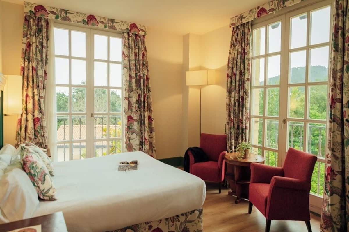 Una habitación de hotel con una cama, dos sillones rojos, una mesita pequeña y ventanas grandes con cortinas estampadas.