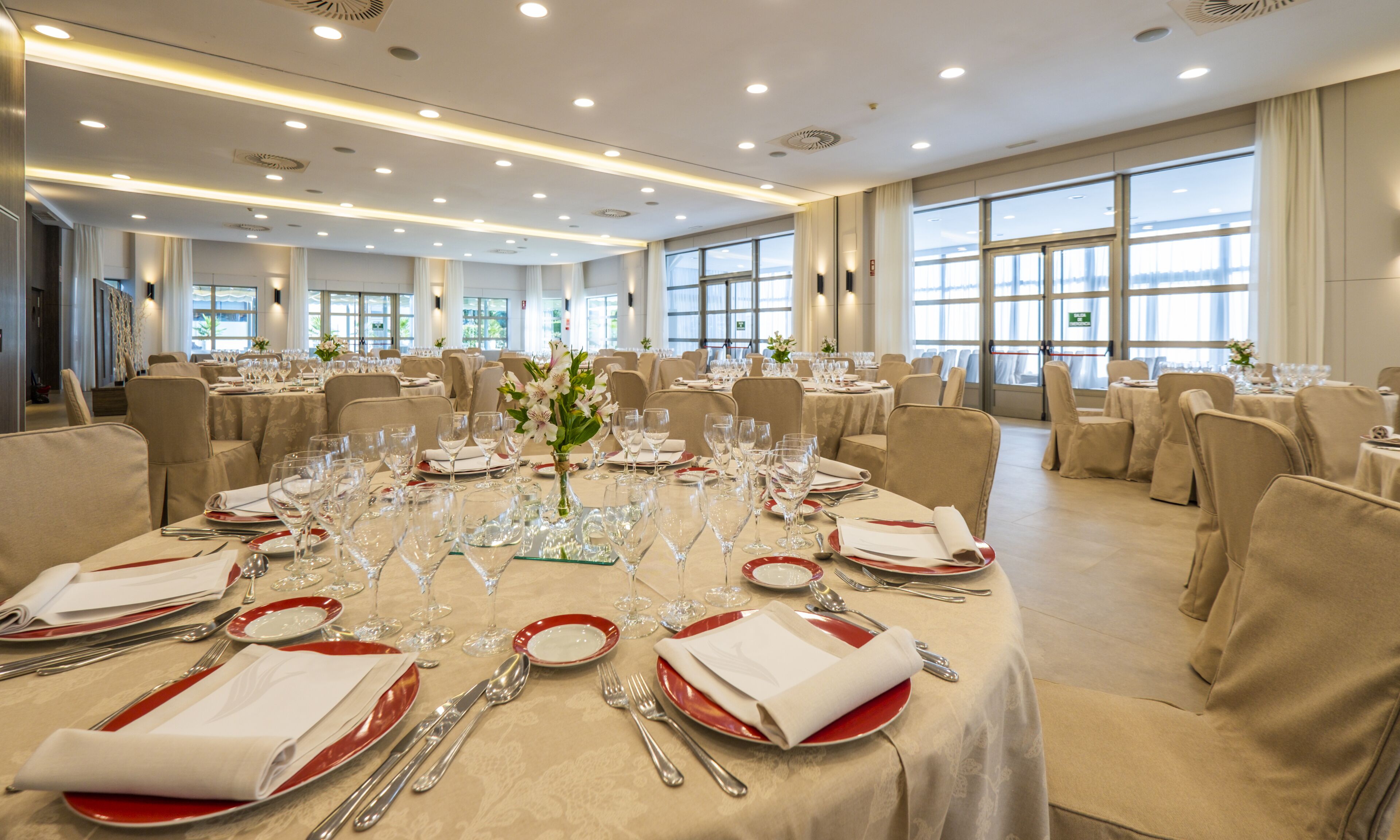 A banquet hall with round tables set for dining, featuring white tablecloths, red plates, and silverware.