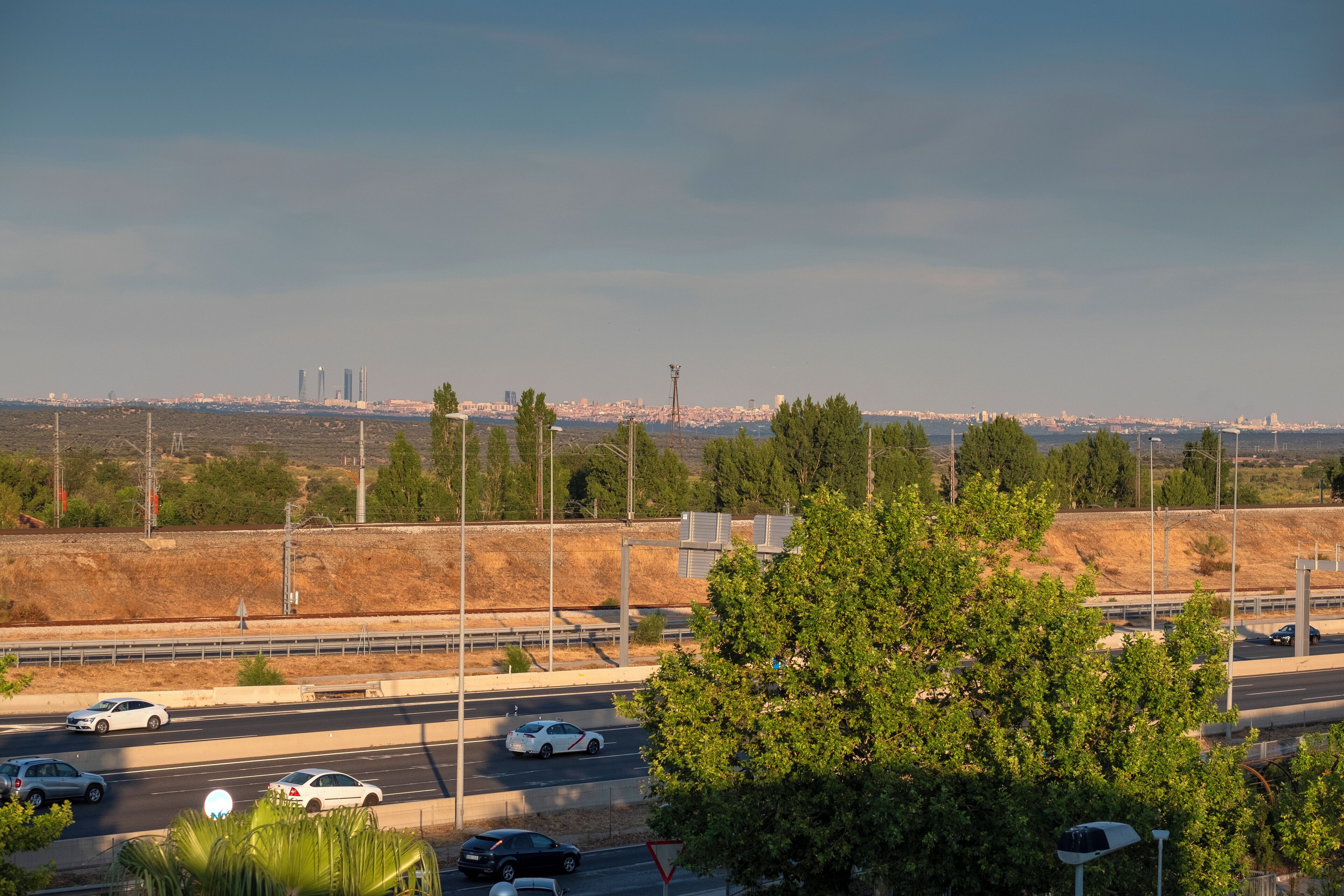 Una autopista con varios carriles y vehículos, rodeada de vegetación y un horizonte urbano distante.