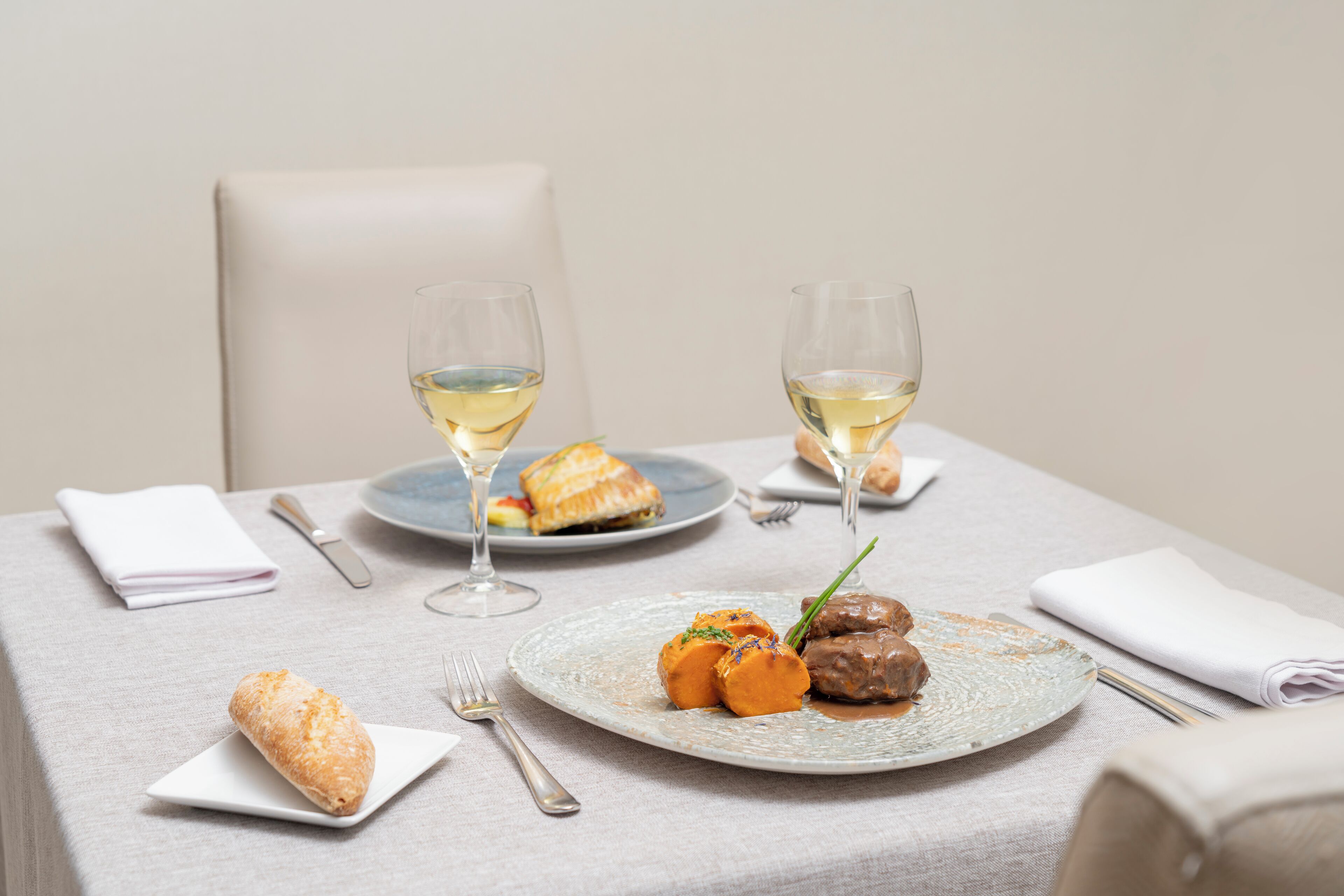Una mesa de comedor con un plato de comida, dos copas de vino blanco, una servilleta doblada y un tenedor.