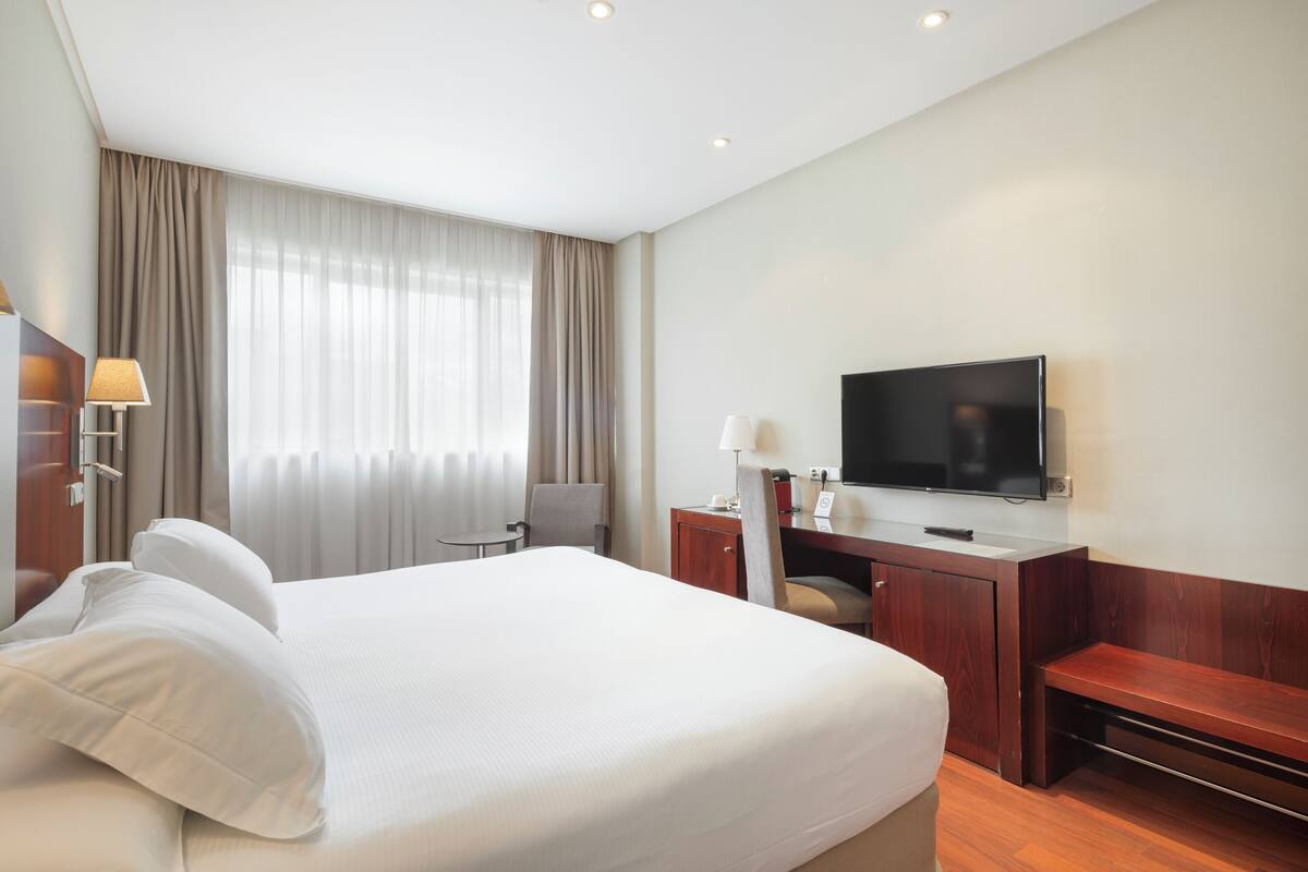 Una habitación de hotel con una cama grande, un escritorio con televisión de pantalla plana, una silla y un banco.
