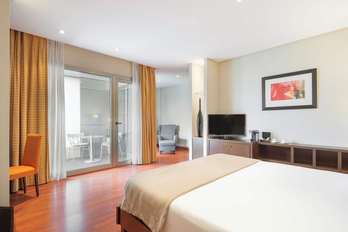 Habitación de hotel con una cama grande, televisión, escritorio y una silla. Posee una puerta de vidrio corredera que da a un balcón.