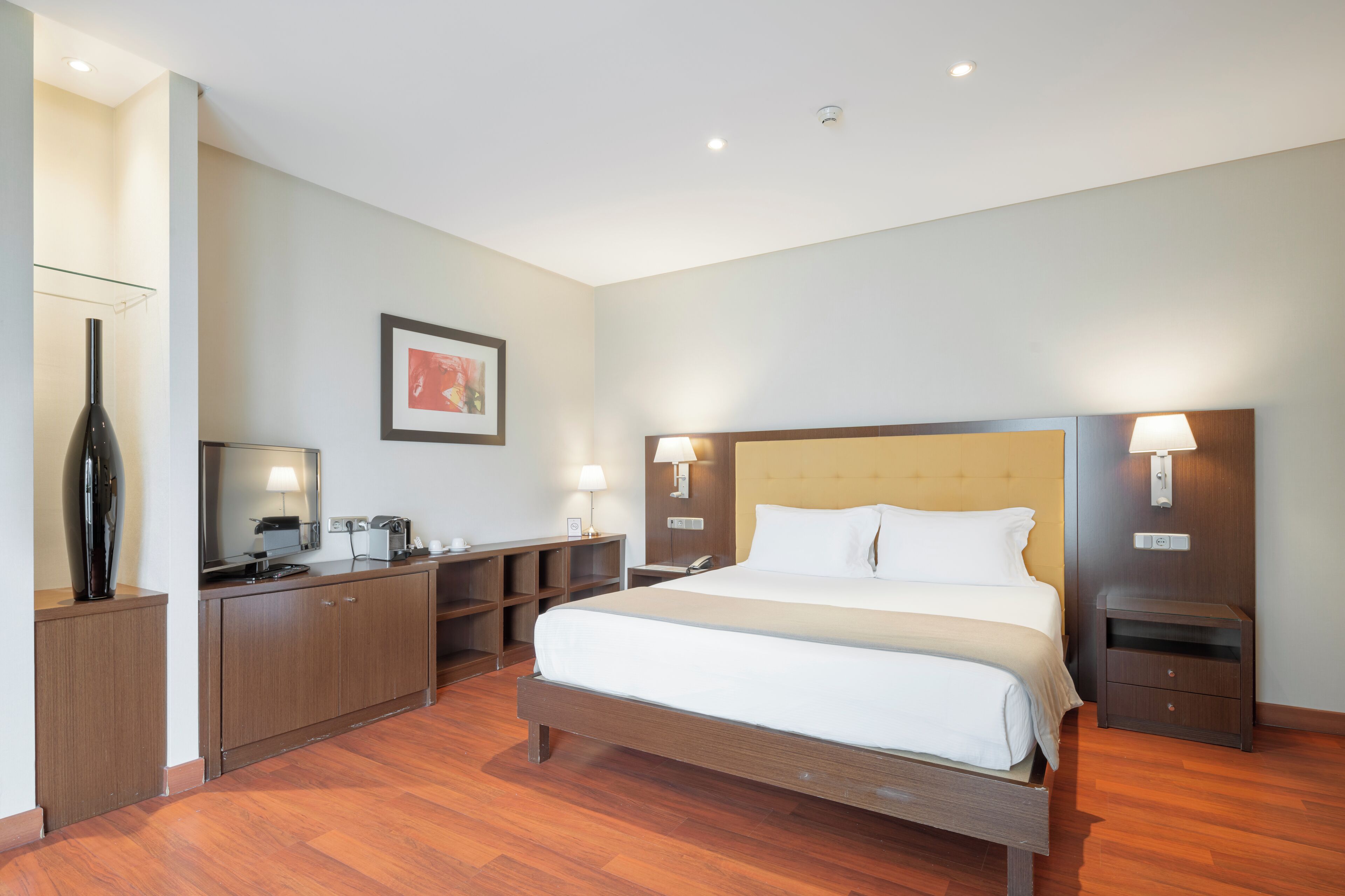 Habitación de hotel con una cama grande, suelo de madera, un escritorio con una silla, una mesita y una mesita de noche.