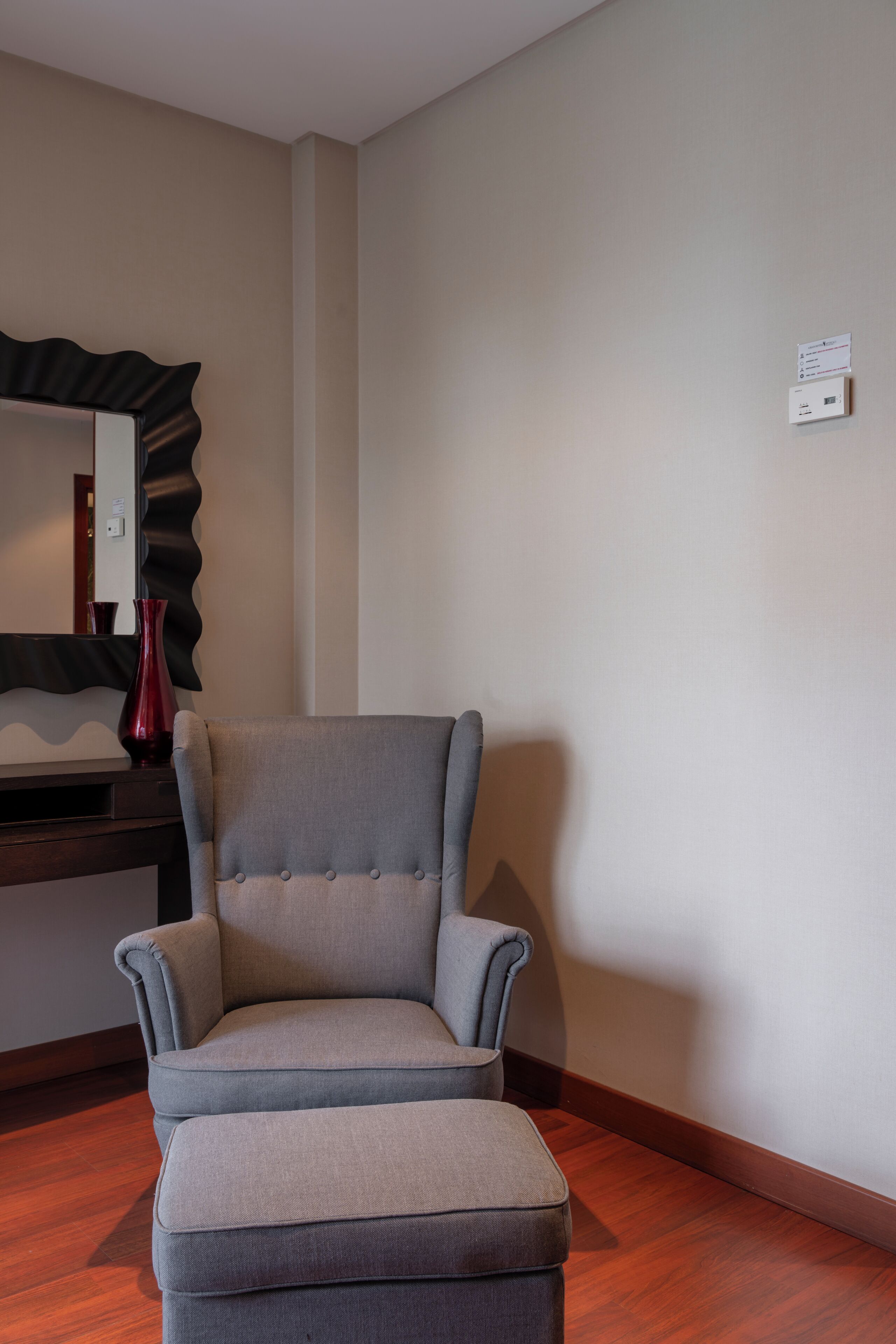 A room with a grey upholstered armchair, a wooden desk, and a red vase.