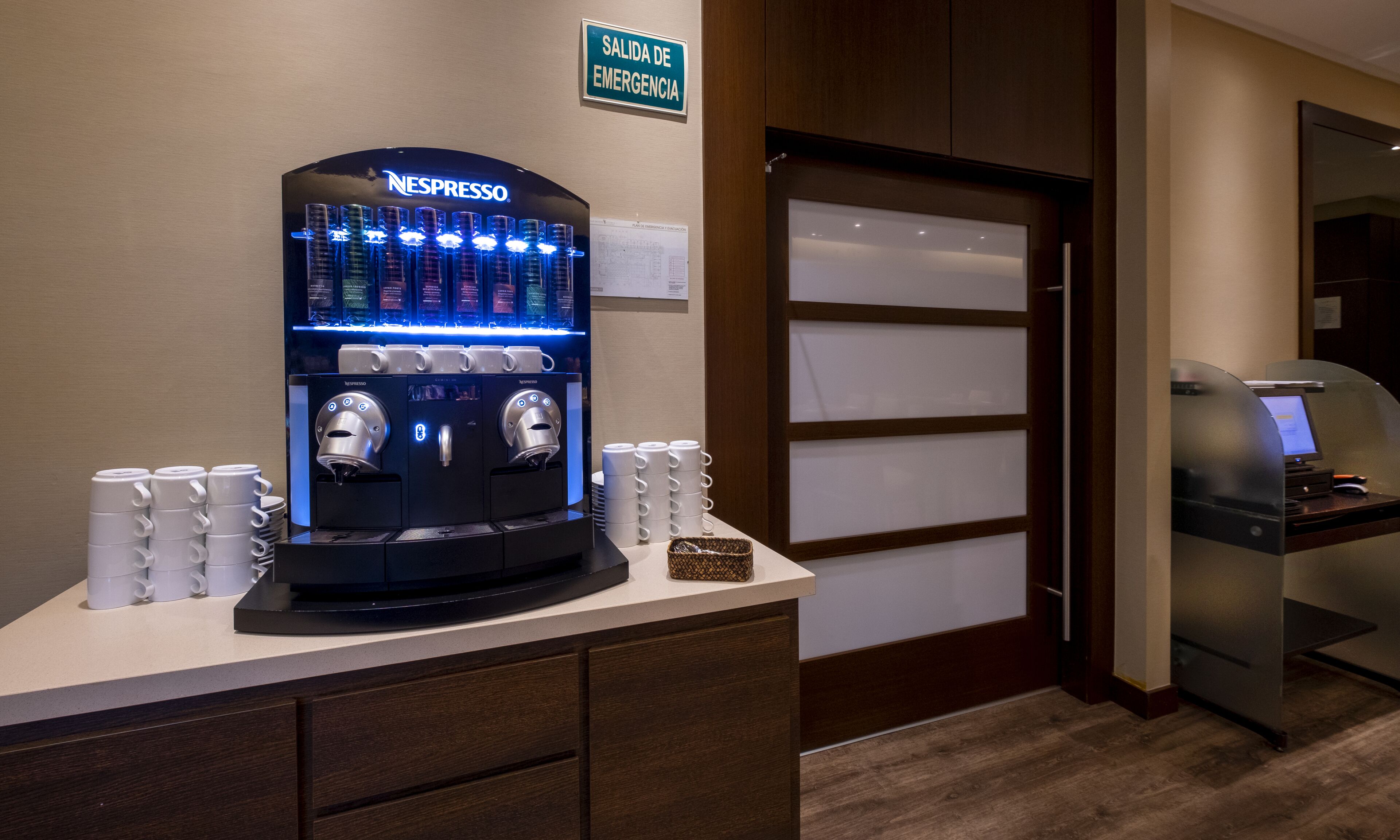 A Nespresso coffee machine with multiple coffee dispensers, a sign indicating an emergency exit, and a glass partition door.