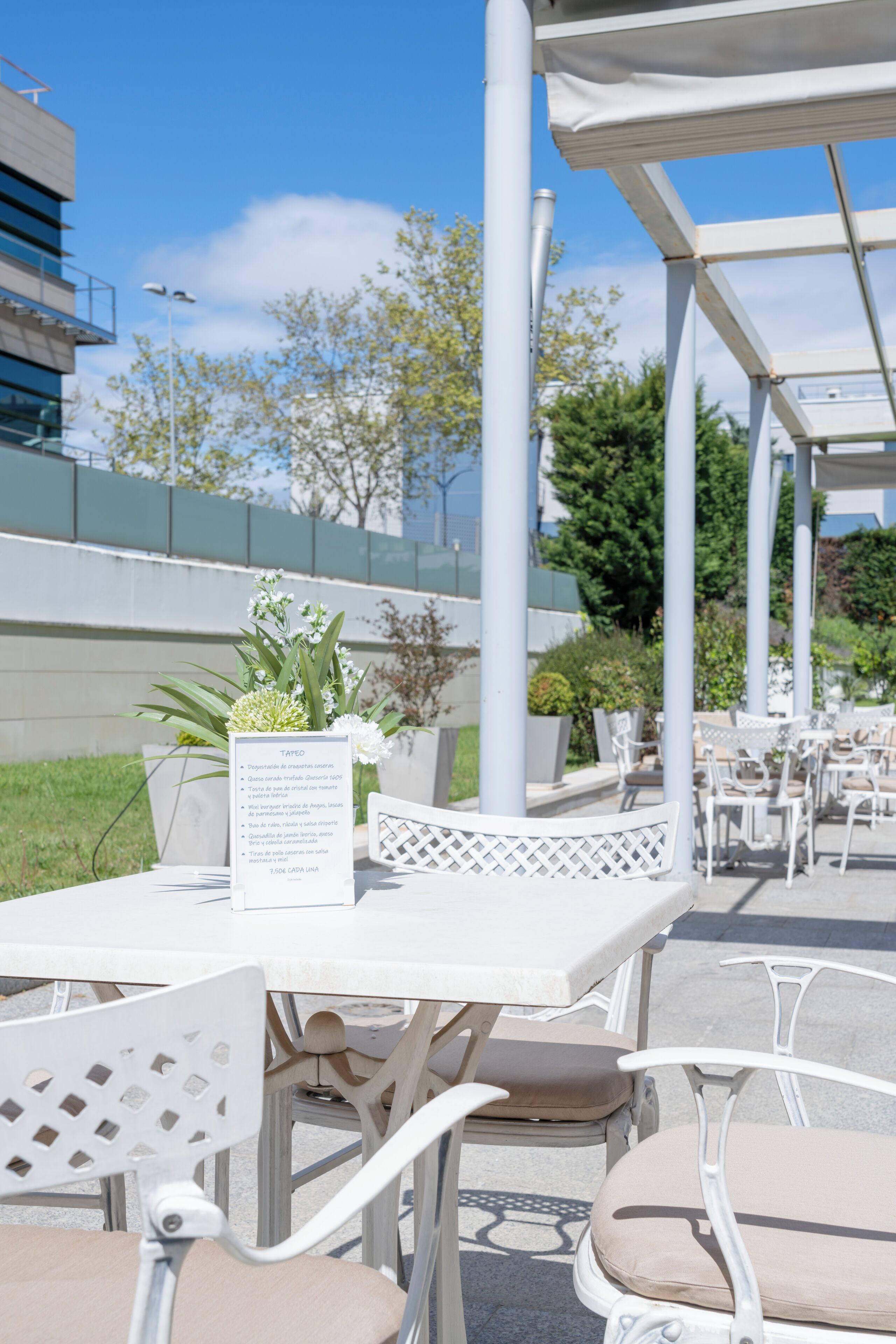 An outdoor seating area with white metal tables and chairs, a pergola structure, and a menu board.