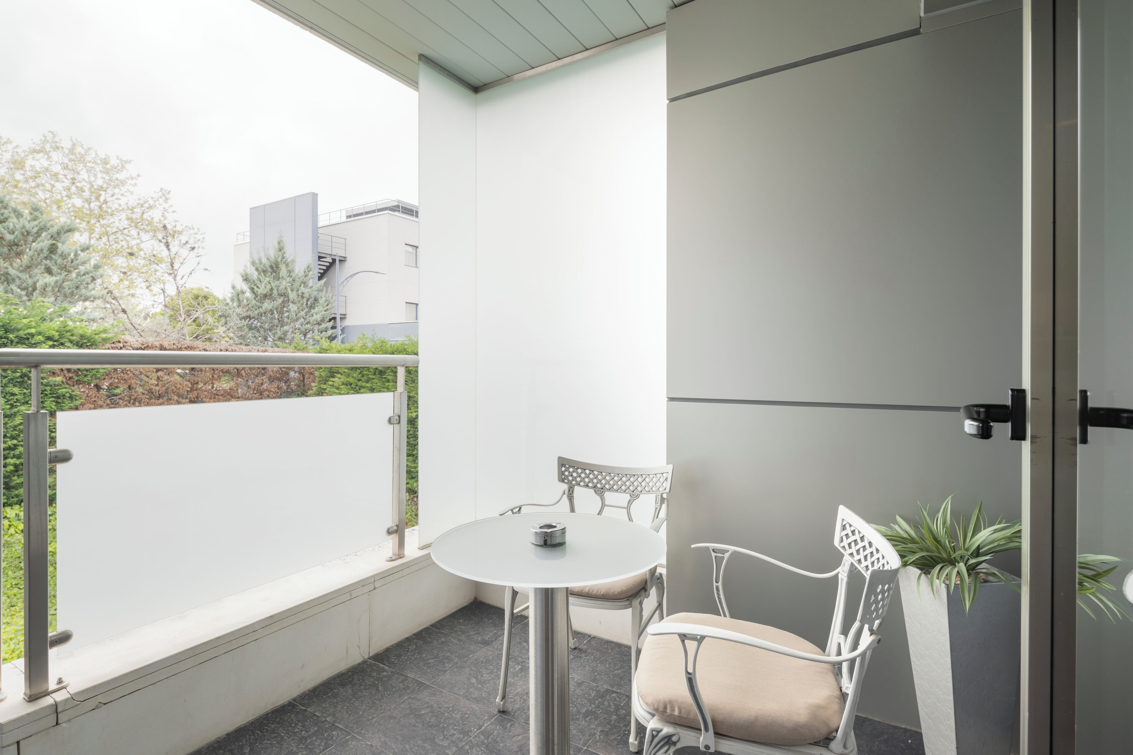 A balcony with a round table and two chairs, a potted plant, and a view of buildings and trees.