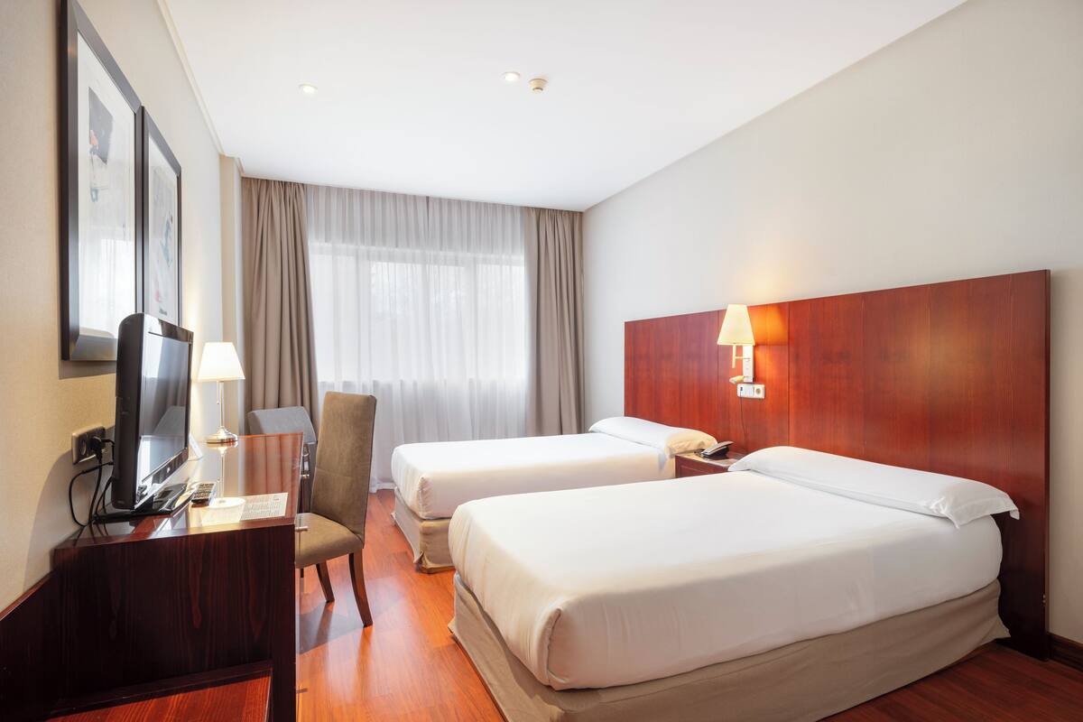Habitación de hotel con dos camas, un escritorio con lámpara, una silla, un televisor y una ventana con cortinas.