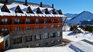 Un edificio de varios pisos con fachada de piedra y balcones de madera, frente a un paisaje montañoso nevado.