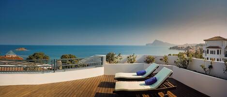 A rooftop terrace with two sun loungers, a wooden deck, and a view of the sea and distant landscape.