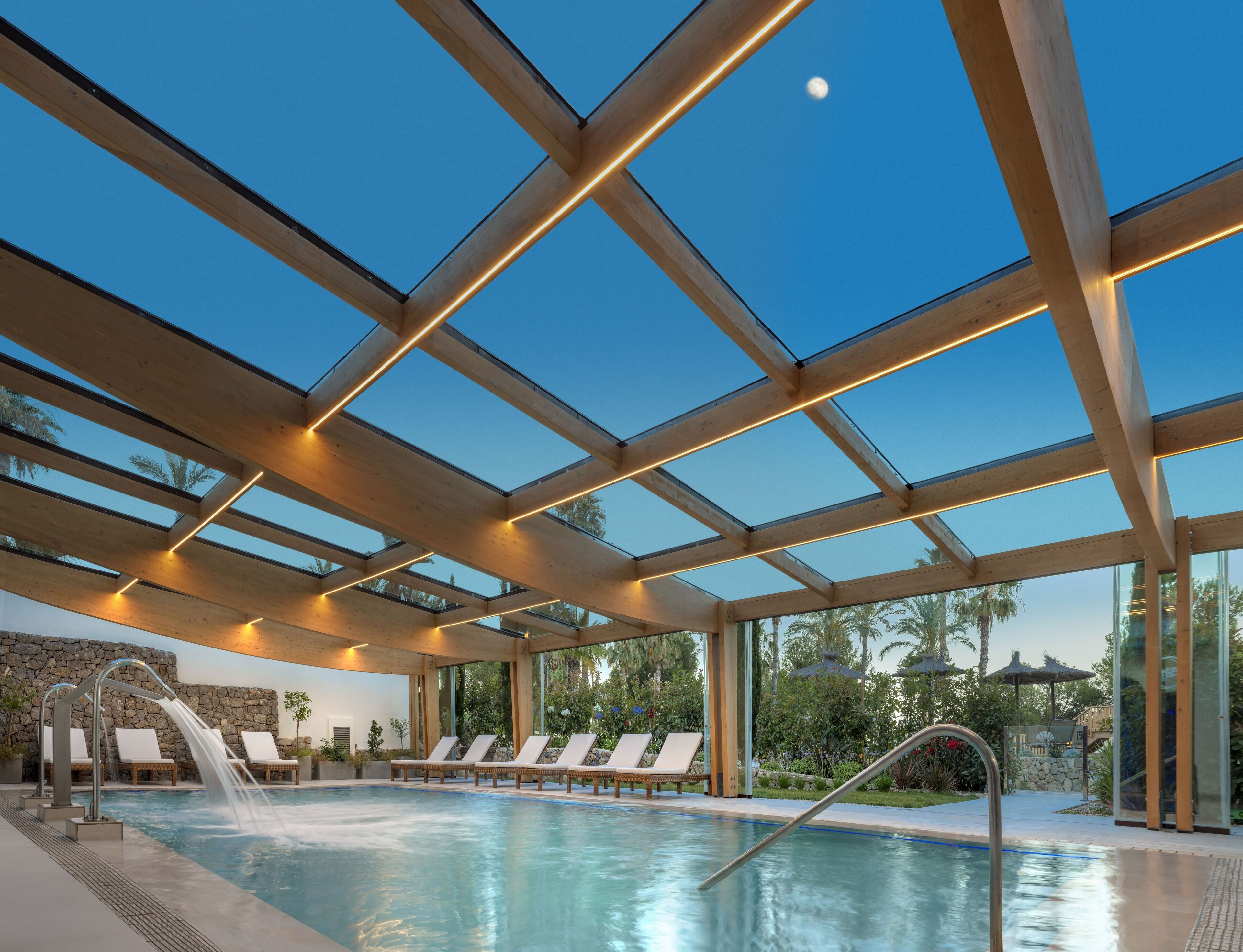 An indoor swimming pool with a glass ceiling, lounge chairs, and a waterfall feature.