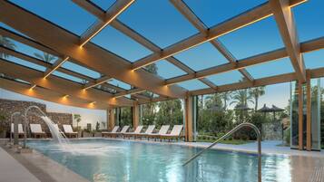 Una piscina interior con techo de vidrio, sillas de descanso y una cascada.