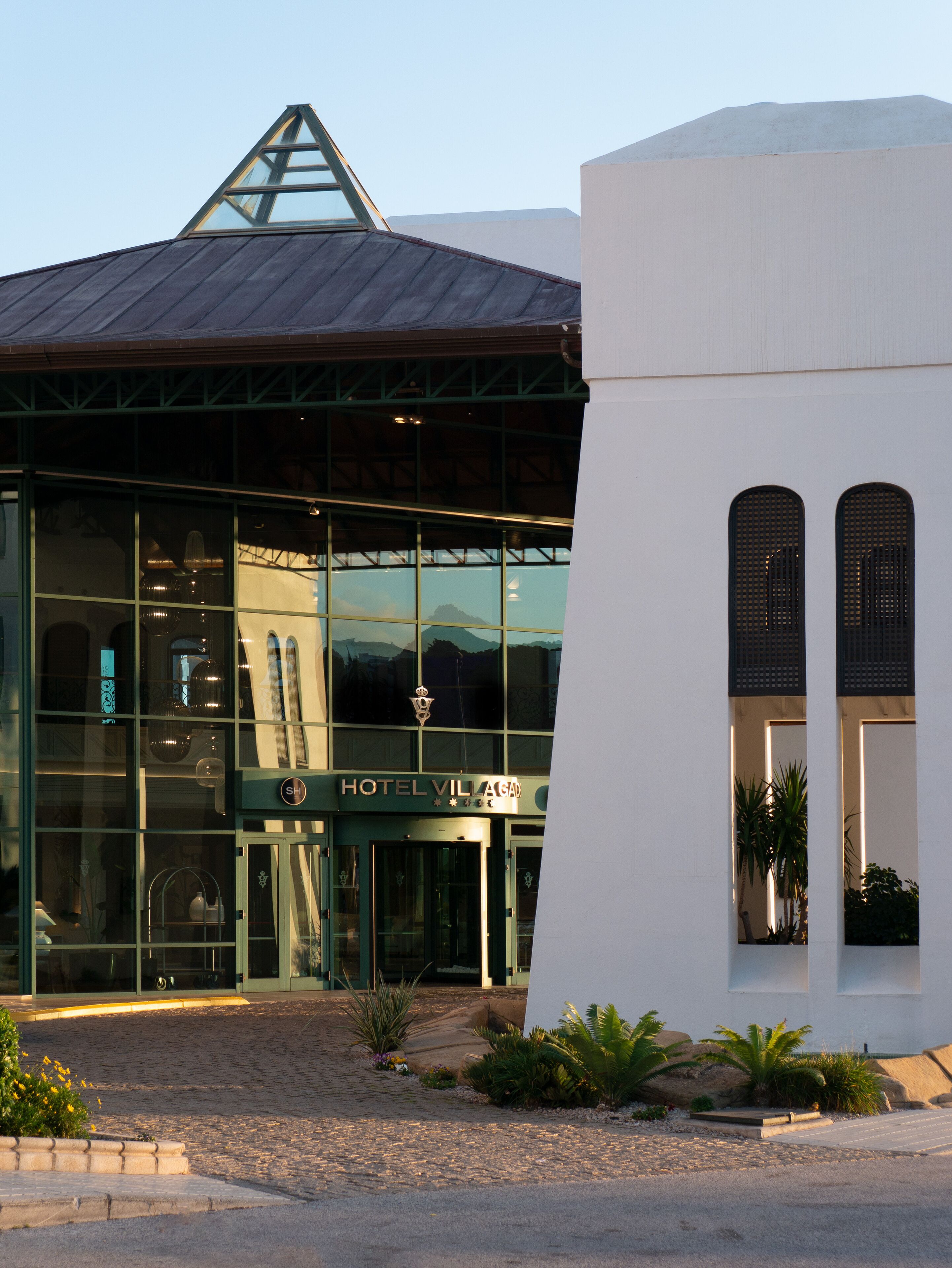A modern hotel with a glass facade, a metal roof, and a sign reading "HOTEL VILLAGE".