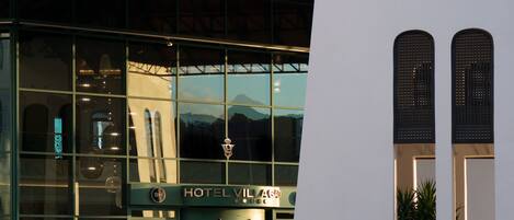 A modern hotel with a glass facade, a metal roof, and a sign reading "HOTEL VILLAGE".