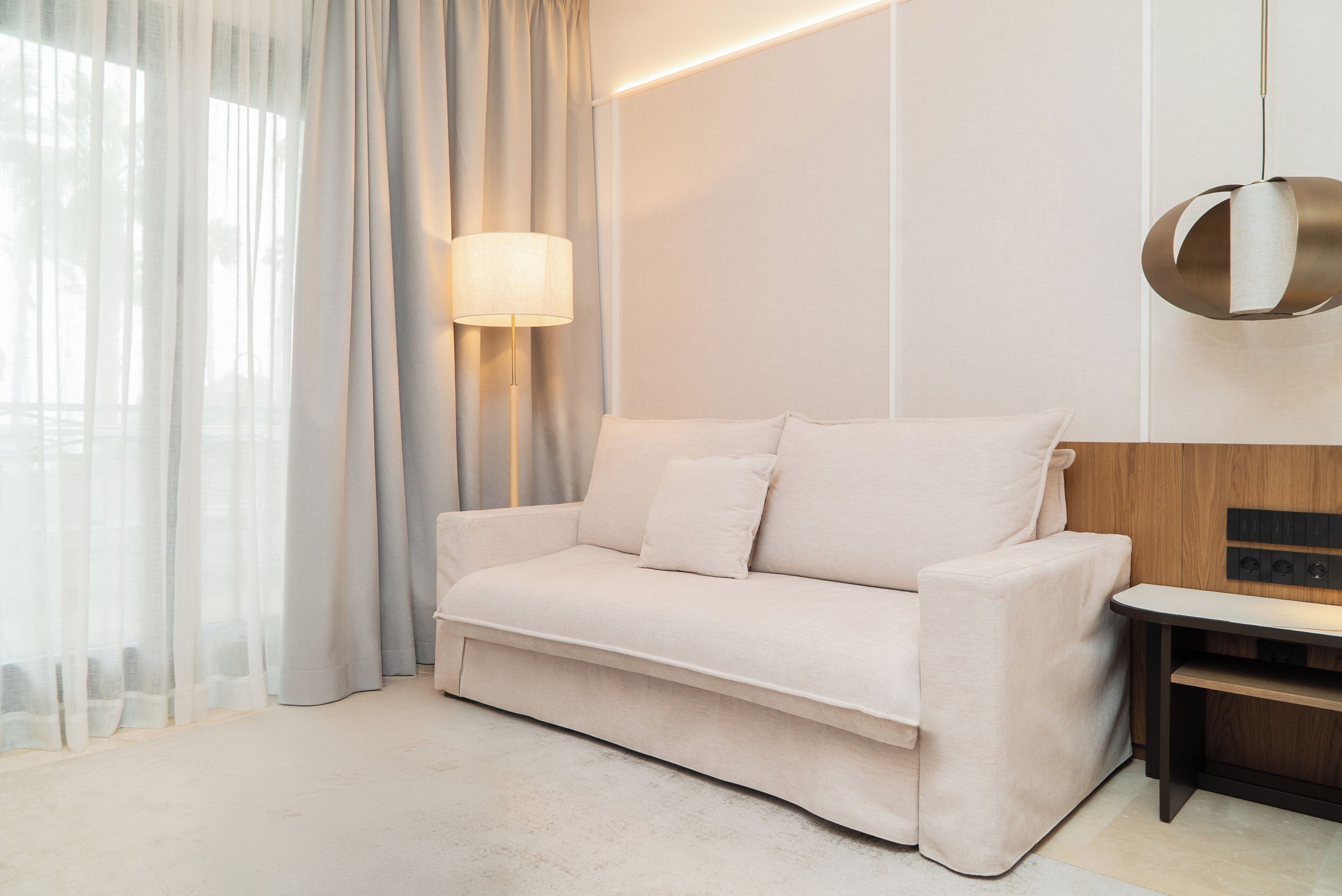 Una habitación de hotel moderna con sofá, lámpara, mesa y vistas a través de cortinas translúcidas.