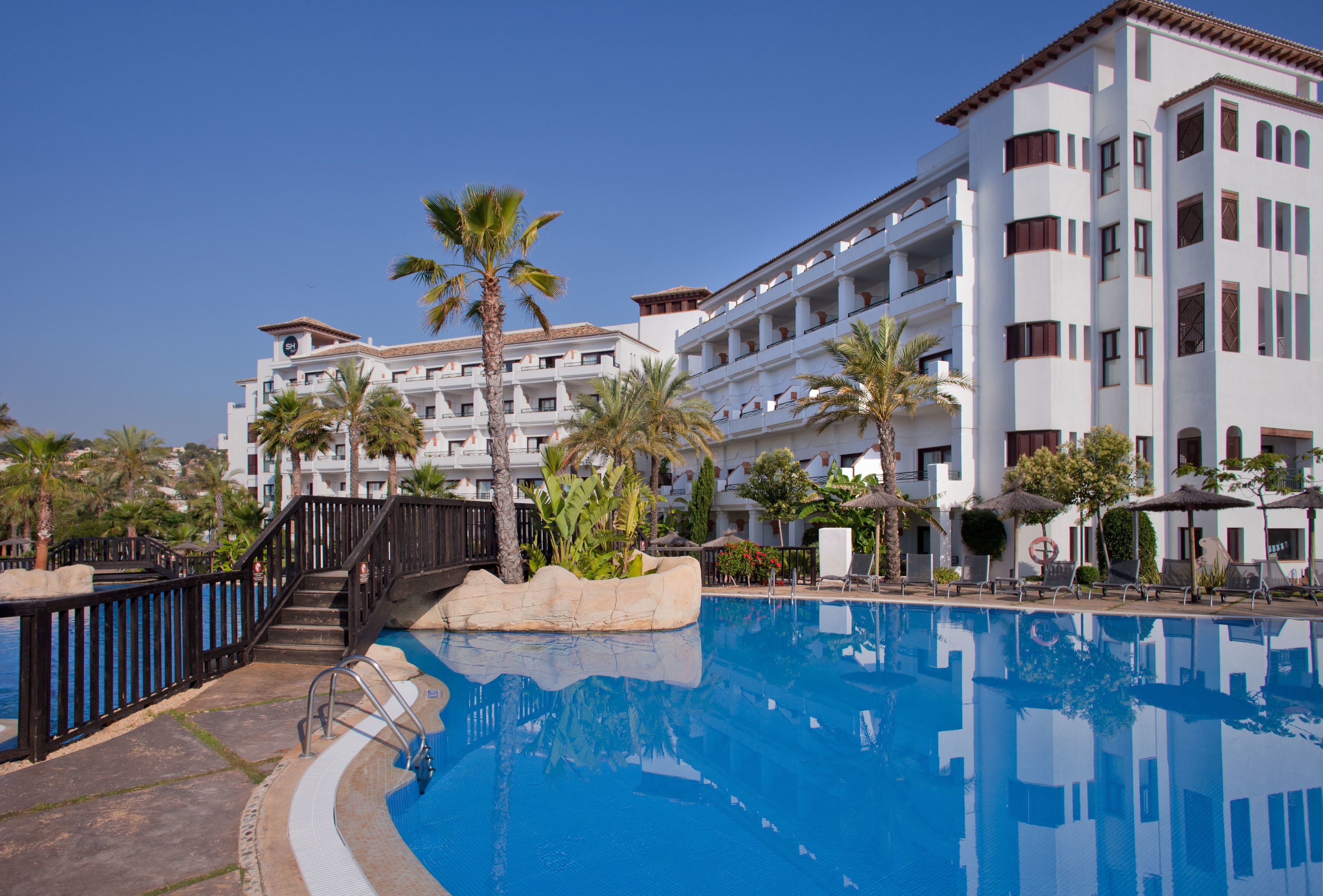 Zona de piscina en un resort con piscina azul cristalina, palmeras y un edificio de varios pisos con balcones.