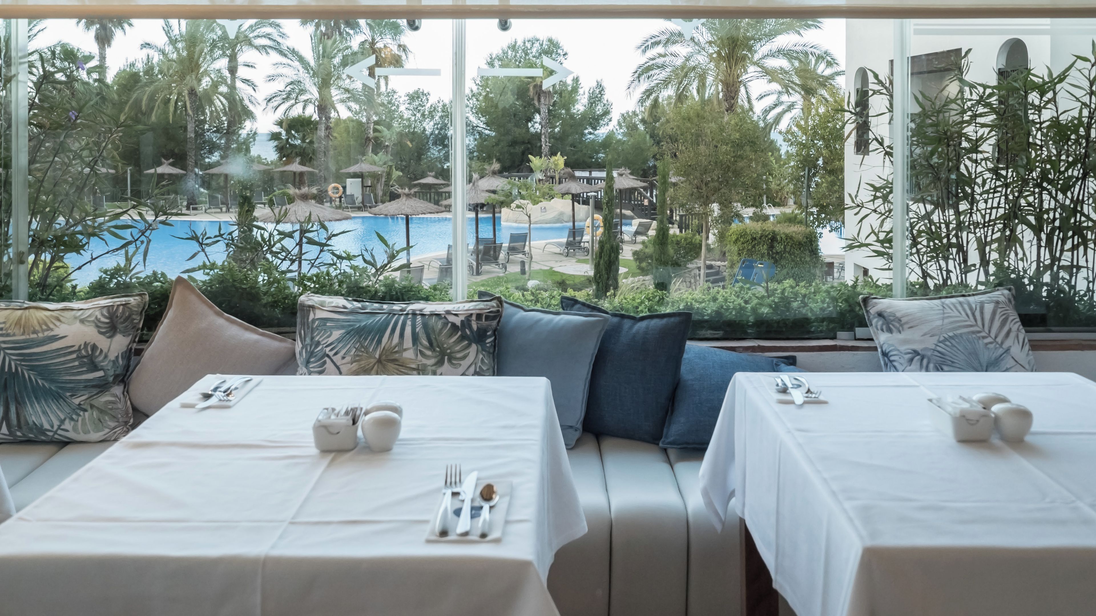 Comedor con mesas preparadas para dos, con vista a una piscina y palmeras.