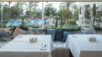 Comedor con mesas preparadas para dos, con vista a una piscina y palmeras.