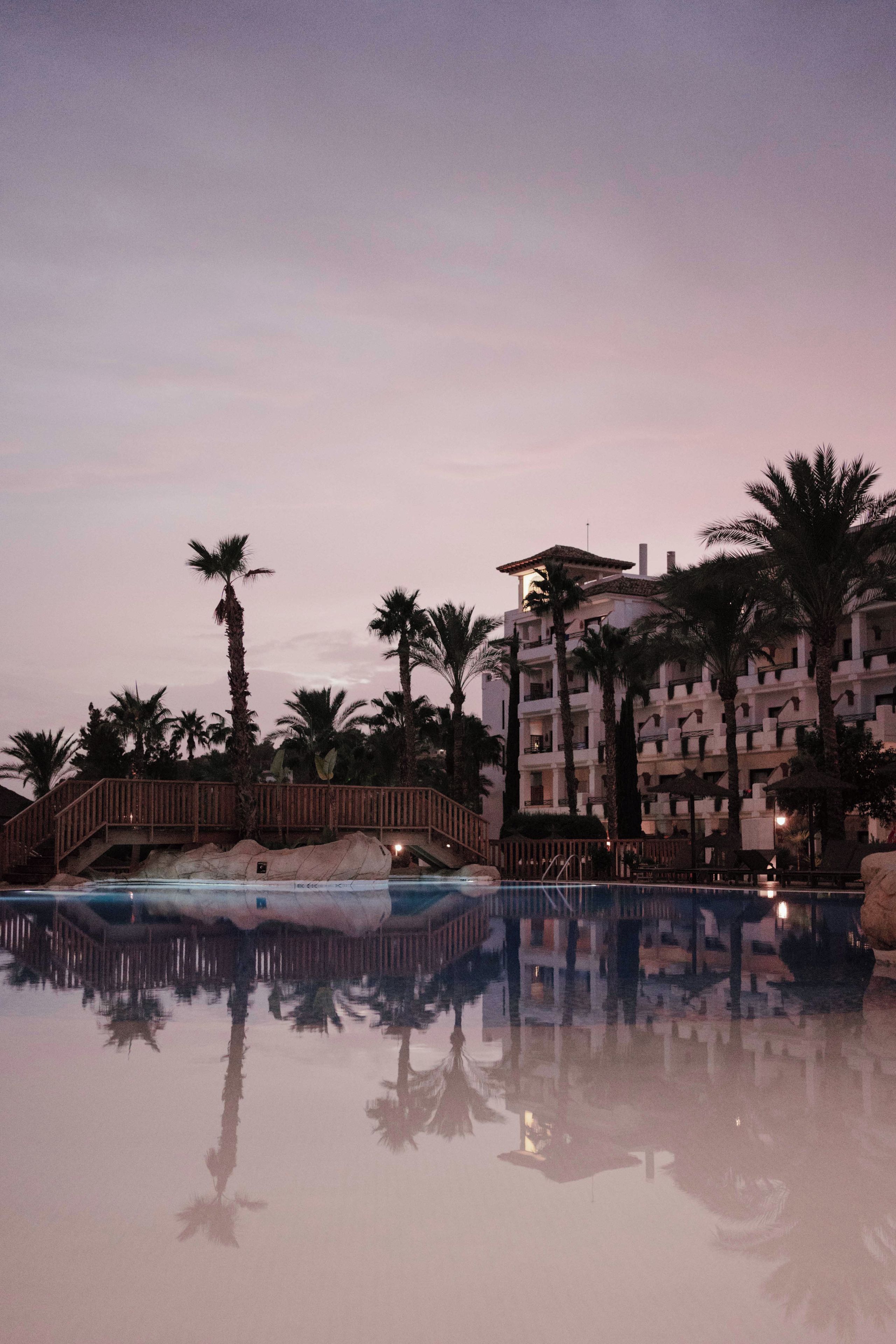 Een resort zwembad met een houten terras en palmbomen, omgeven door een meer verdiepingen tellend gebouw met balkons.