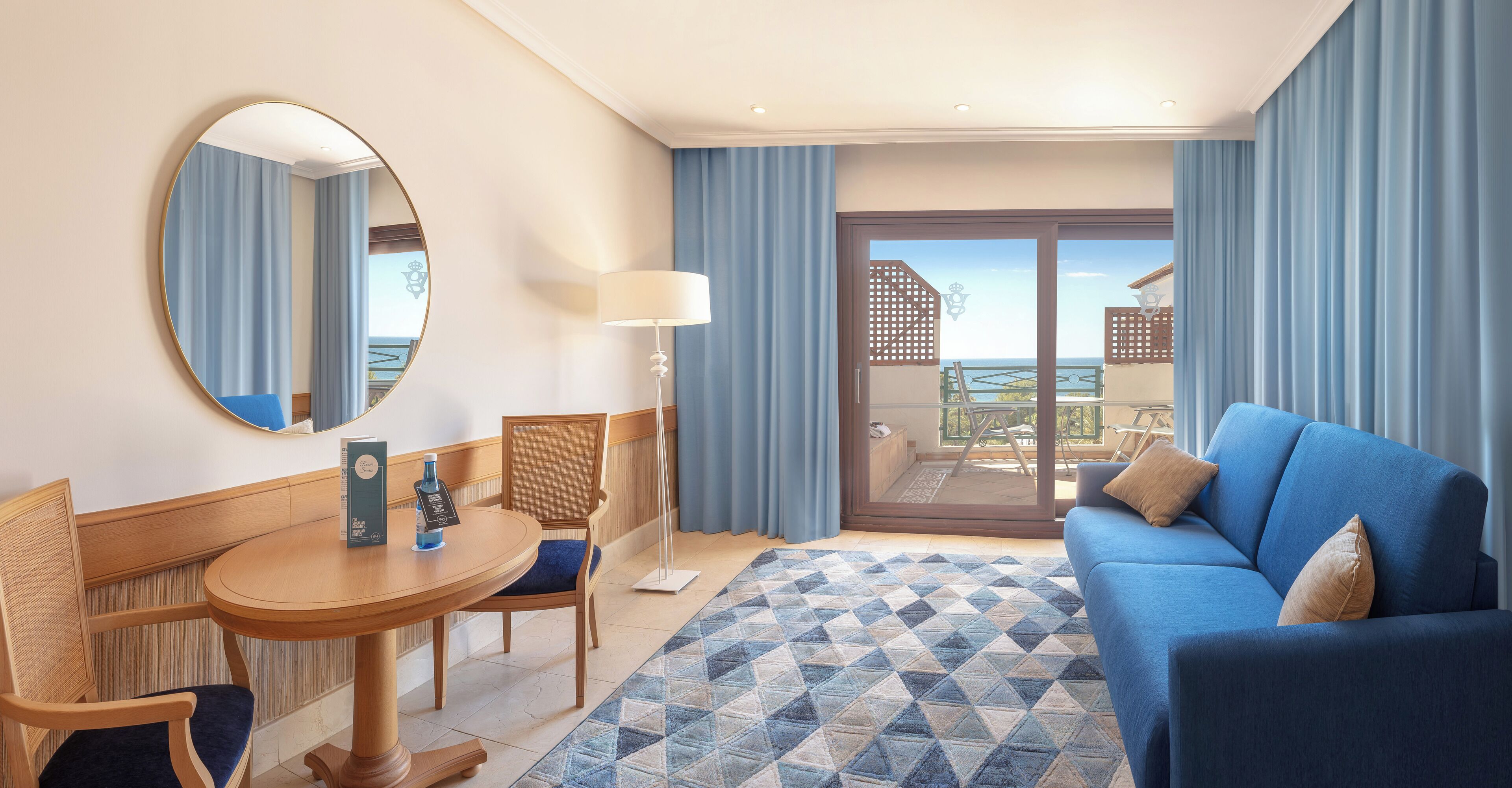A hotel room with a blue sofa, a round wooden table, a chair, a large mirror, and a view of the sea through a sliding glass door.