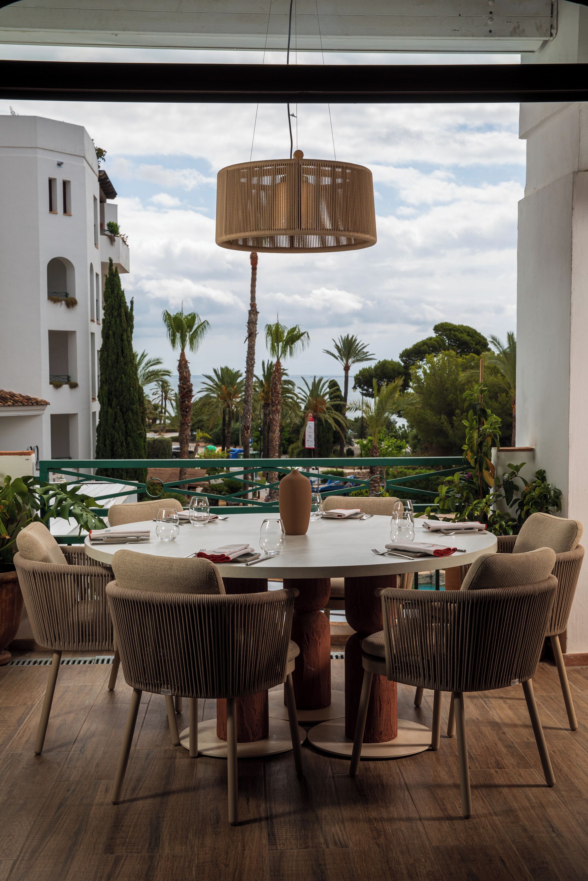 A round dining table with six chairs, set for a meal, overlooking a pool and palm trees.