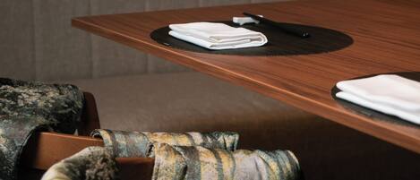 A wooden table with a black placemat, a folded white napkin, and a black pen. A grey upholstered bench is in the background.