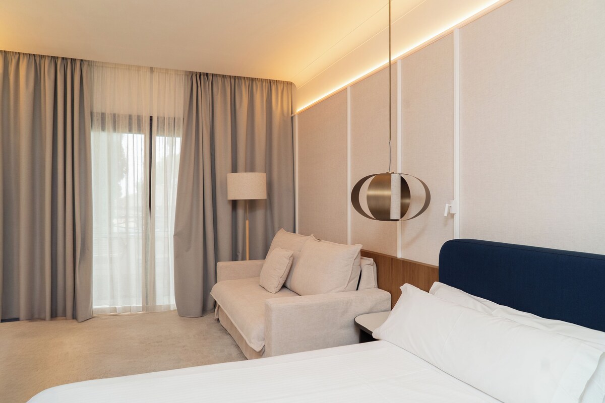 A hotel room with a bed, a sofa, a lamp, and a wall-mounted light fixture.