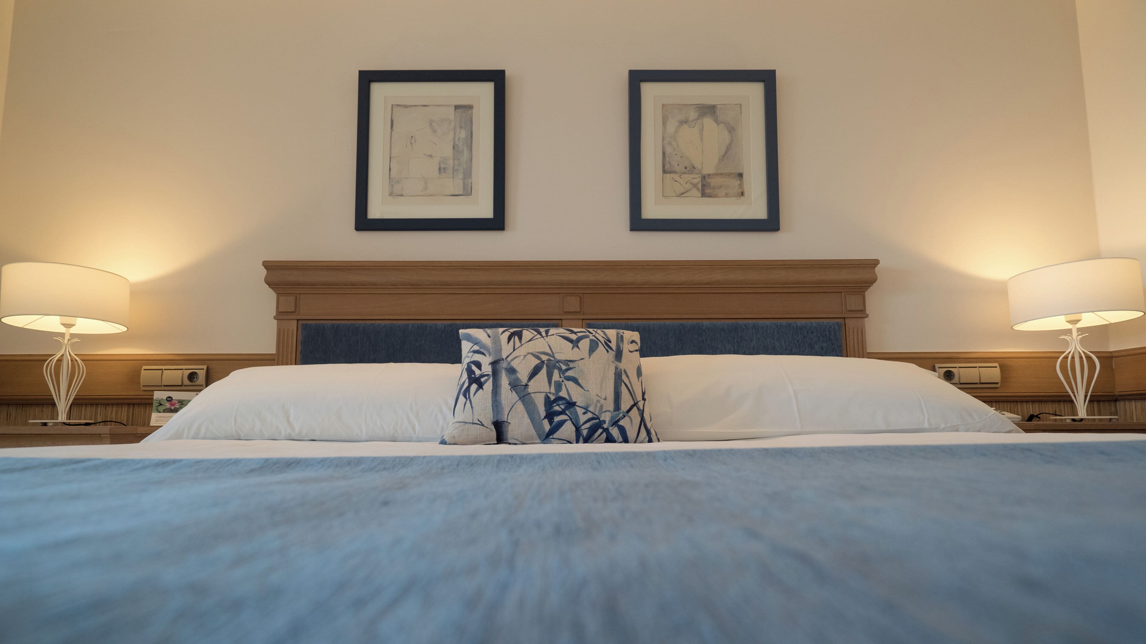A hotel room with a large bed, two bedside lamps, and two framed artworks on the wall.