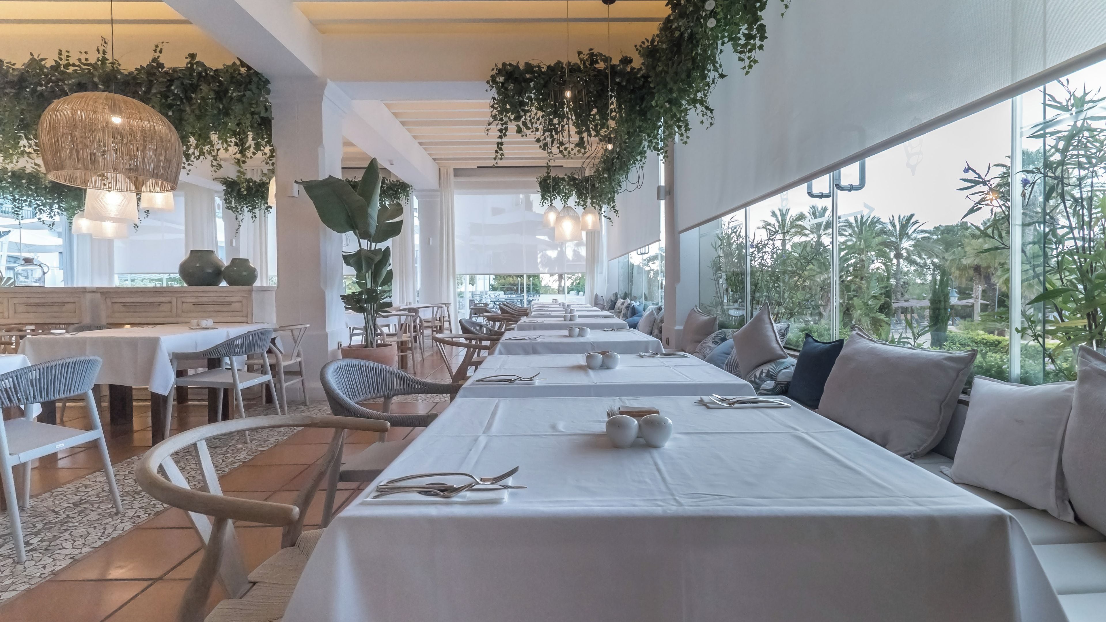 Un restaurant avec de longues tables, des chaises, des plantes suspendues et de grandes fenêtres.