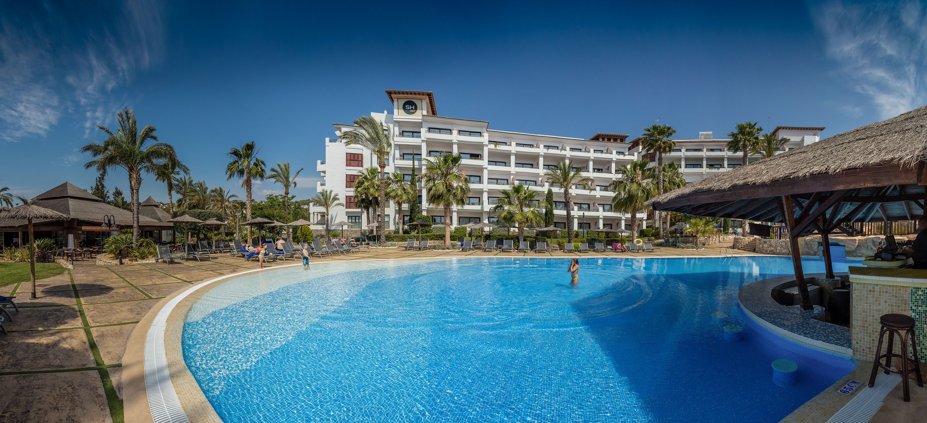 A large swimming pool with lounge chairs and palm trees, surrounded by a hotel building with multiple balconies.