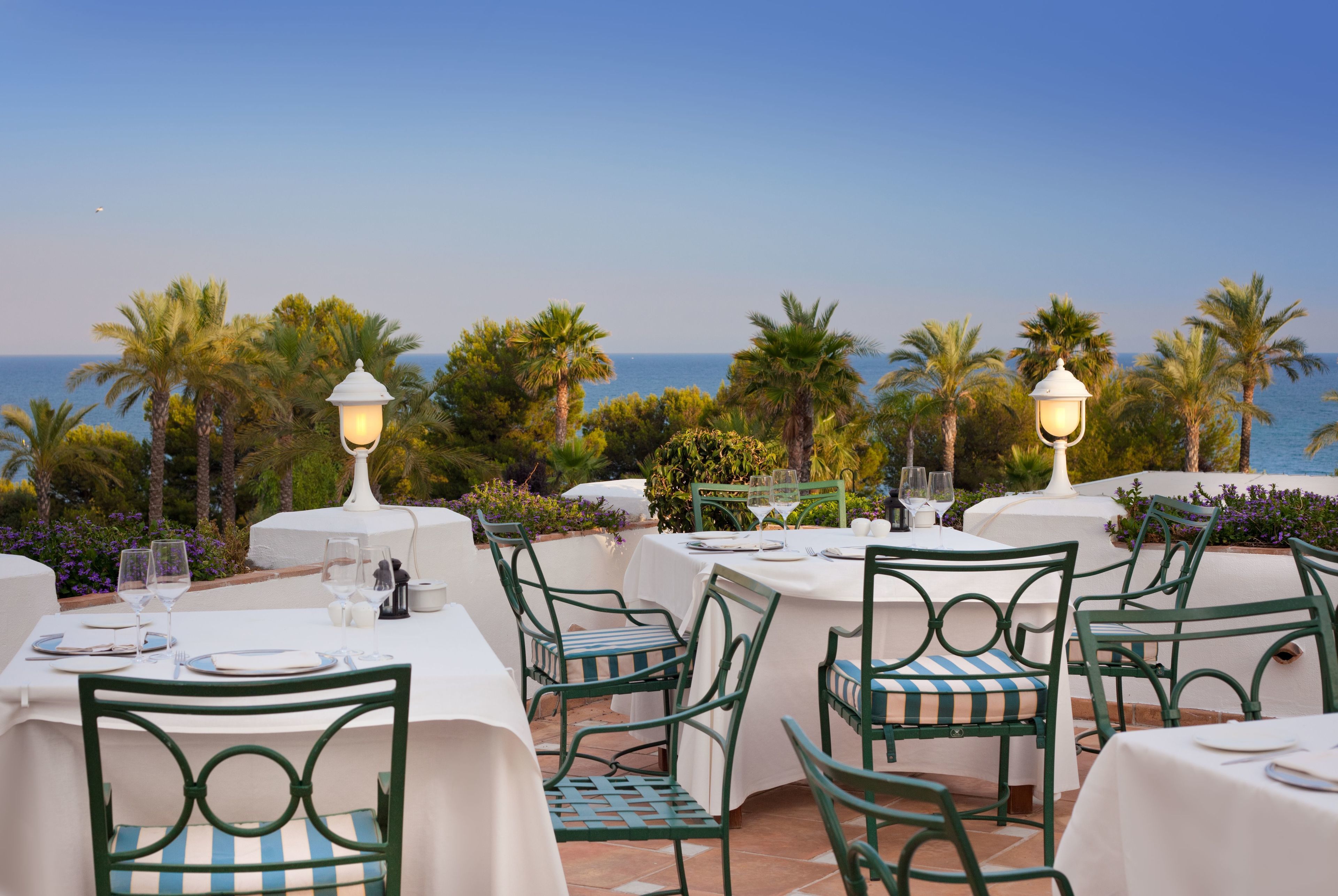 An outdoor dining area with tables and chairs, overlooking a body of water with palm trees in the background.