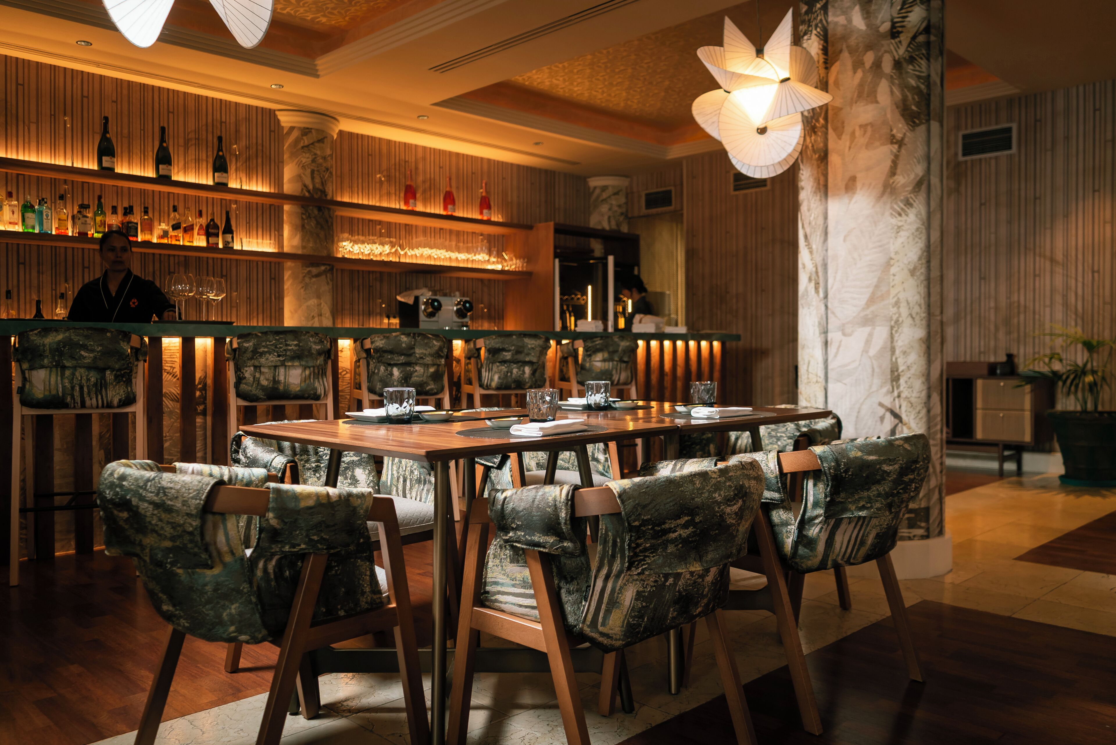 Una zona de bar con un cantinero, una mesa de madera larga con sillas, y estantes con botellas y copas.