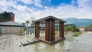 Um pavilhão moderno com estrutura de vidro, rodeado por um espelho d'água e um cenário montanhoso.