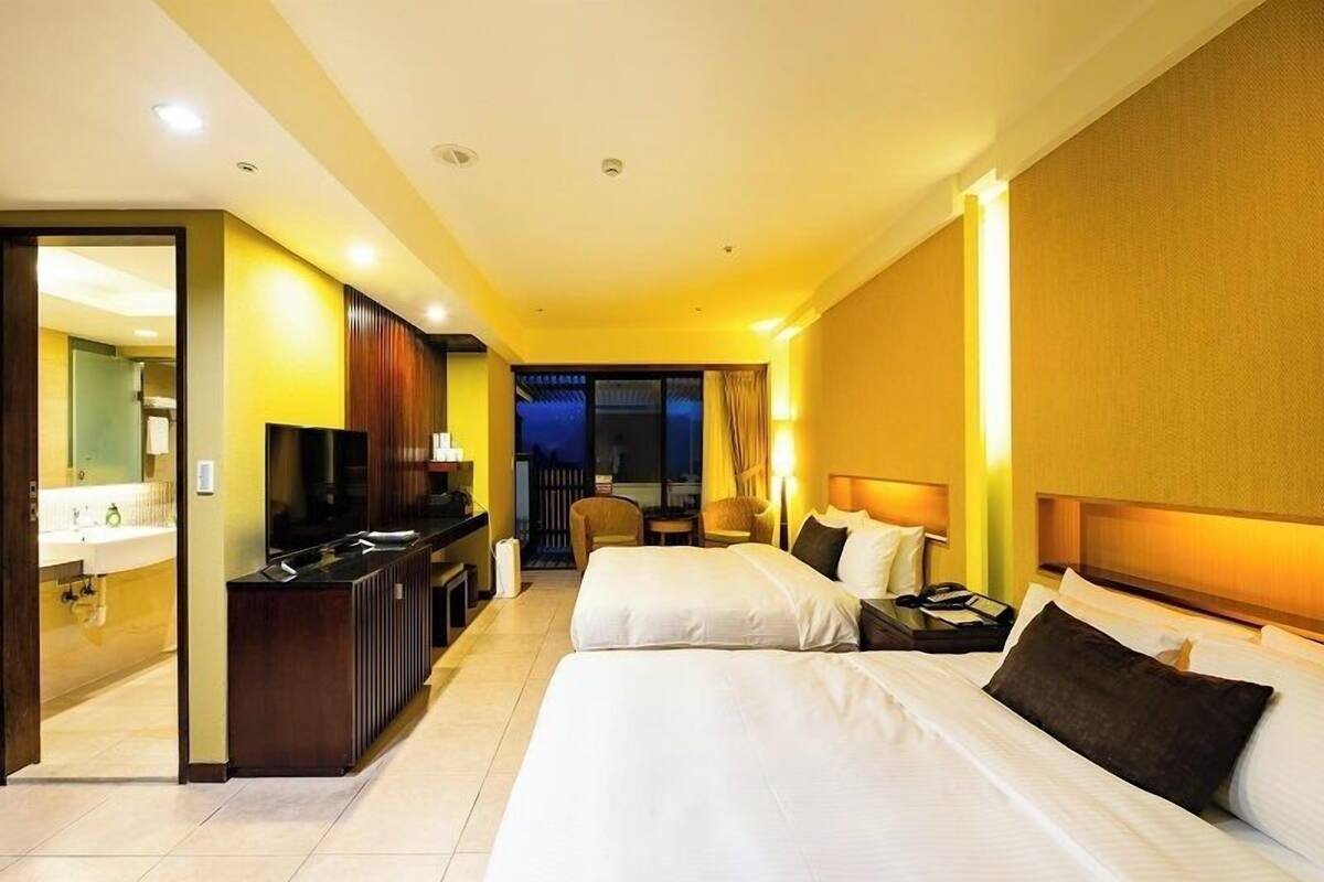 Habitación de hotel con dos camas, un escritorio con televisor y un baño visible a través de una puerta abierta.