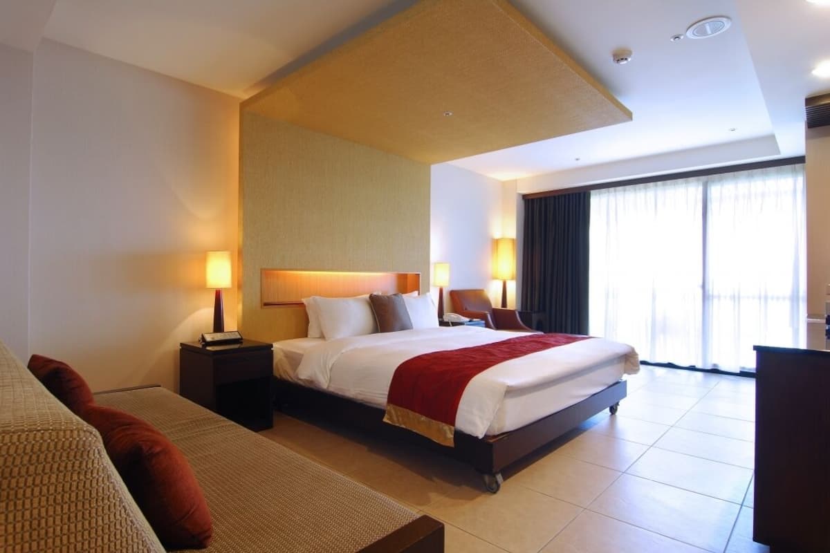 Habitación de hotel con una cama grande, dos mesitas de noche con lámparas, un sofá y un amplio ventanal con cortinas.