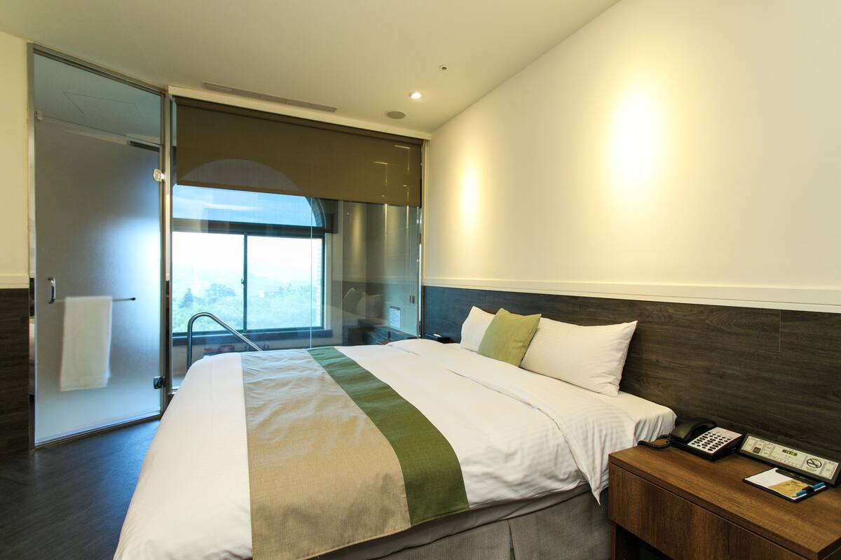 Habitación de hotel moderna con una cama grande, una mesita de noche de madera, un teléfono y vistas al exterior a través de un ventanal.