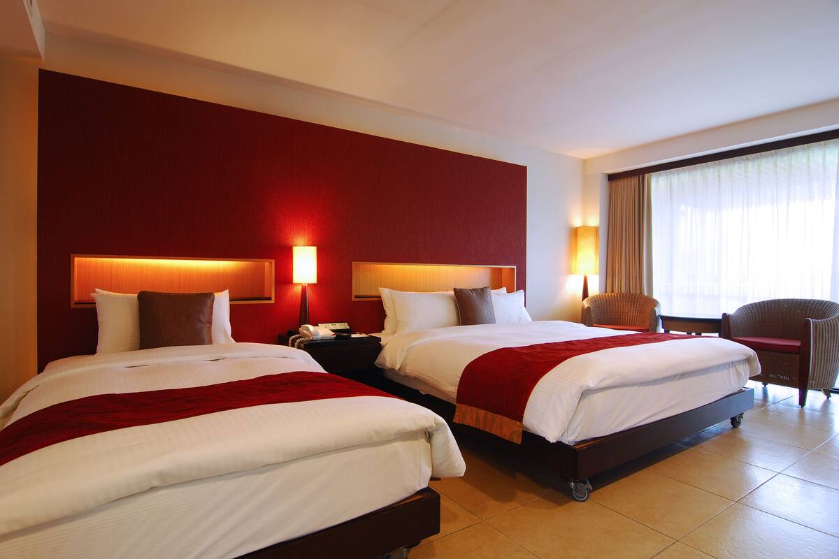 Habitación de hotel con dos camas, una mesita de noche con teléfono, una silla y un amplio ventanal con cortinas.