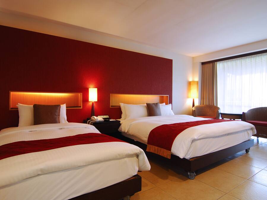 Habitación de hotel con dos camas, una mesita de noche con teléfono, una silla y un amplio ventanal con cortinas.