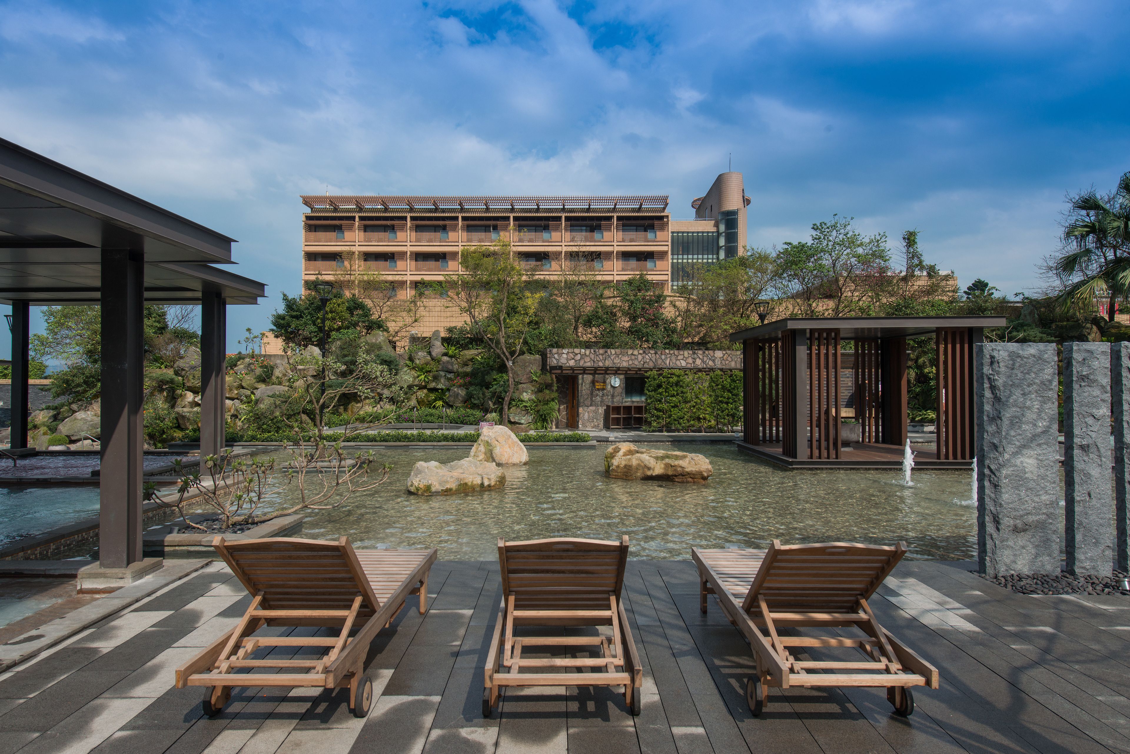 A serene outdoor area with a pool, wooden lounge chairs, and a building in the background.